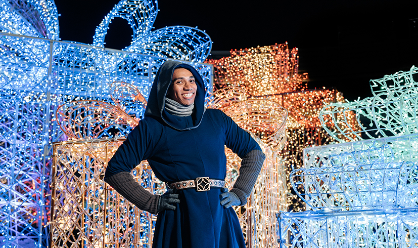 smiling elf in a robe in front of huge lightened gift box sculptures at the Enchant Christmas village