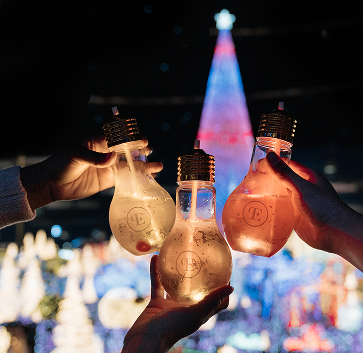 hands holding light bulb glasses with drinks at the enchant christmas village